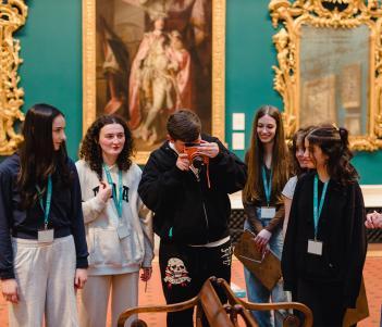 A teenage boy takes a photo on a bright orange camera in a grand gallery space. A group of five teenage girls watch on.
