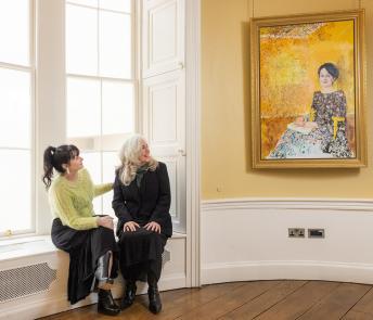 Photograph of two women, one wearing black, the other wearing a green top, sitting in a yellow room looking at a painting