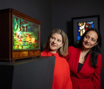 Photograph of two women, both wearing red dresses, looking around a corner at a piece of decorative stained glass.