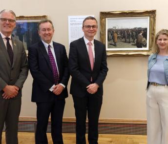 Three men wearing dark suits and a woman wearing white trousers and a blue top stand in front of a painting.