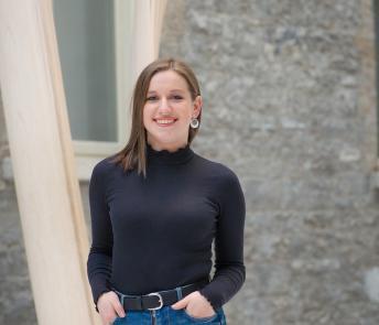 Sarah stands in the Gallery Courtyard. She is smiling, with her hands in her pockets.