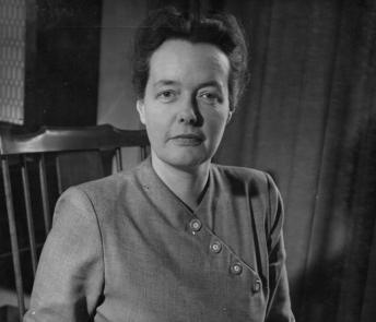 Photograph of Bea Orpen, sitting on a chair, looking at the camera with a book of her artwork open in front of her.