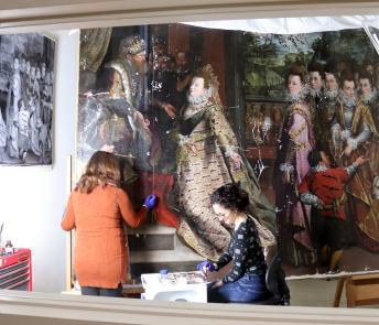 View through a window into a conservation studio where two women work on a large oil painting