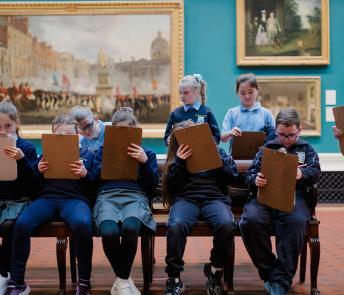 A group of school children in uniform draw on clipboards in a grand gallery filled with paintings.