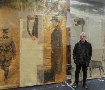 Artist Hughie O'Donoghue standing in his studio in front of two large artworks depicting Michael Collins and Wuffa.
