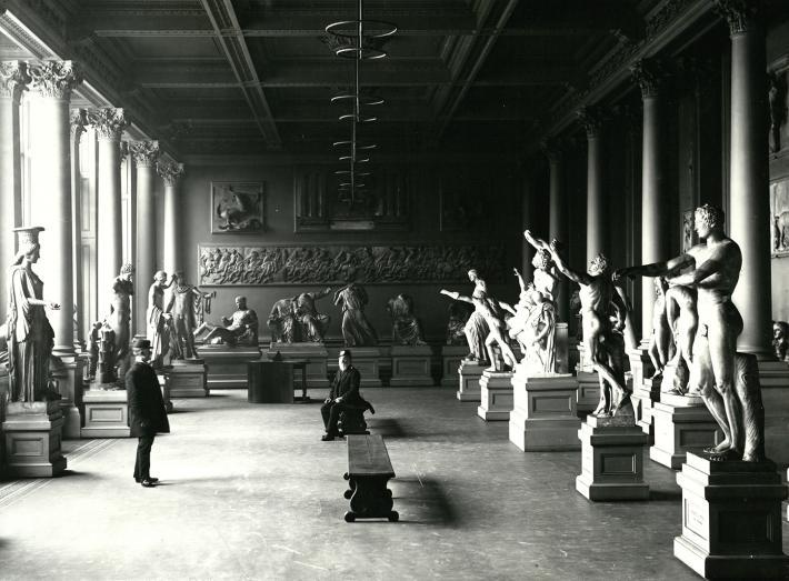 A black and white photograph of a room filled with plaster casts of sculptures. There are two me in the room - one sits on a bench in the middle of the room, the other stands on the left, facing the seated man.
