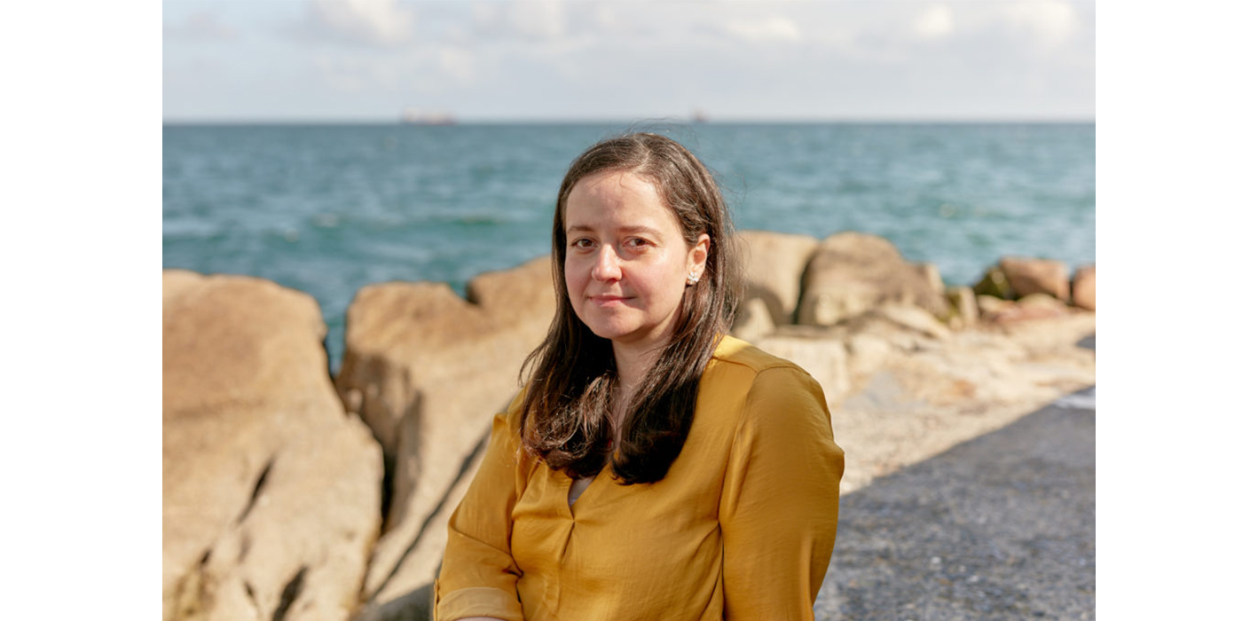 Portrait of a woman with brown hair and wearing a mustard coloured top, pictured sitting on the coast with the sea in the background.